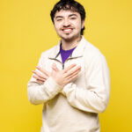 A young man with short, curly dark hair stands against a bright yellow background. He is wearing a light-colored, long-sleeve shirt and is smiling while making a hand gesture with both hands, crossing them in front of his chest. His expression is friendly and approachable.
