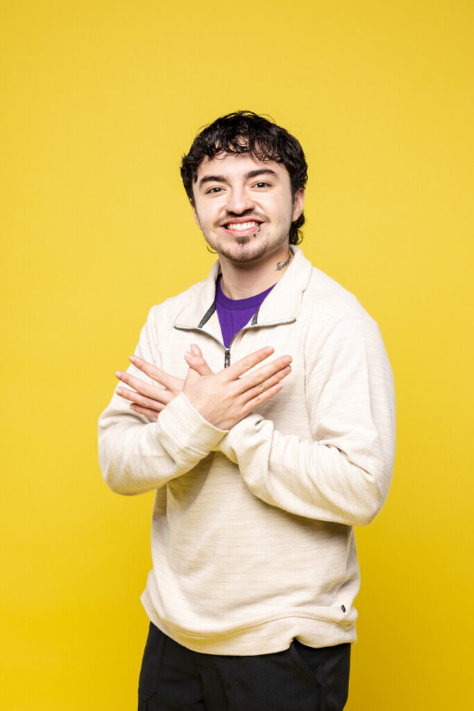 A young man with short, curly dark hair stands against a bright yellow background. He is wearing a light-colored, long-sleeve shirt and is smiling while making a hand gesture with both hands, crossing them in front of his chest. His expression is friendly and approachable.