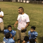 Man in a white shirt stands a football field. He is talking to a kids team in light blue uniforms.