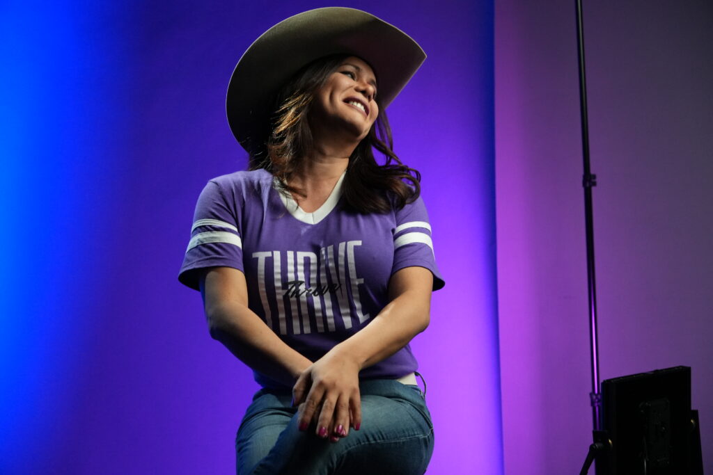 A person wearing a wide-brimmed hat and a purple “Thrive” T-shirt sits on a stool, smiling and looking off to the side against a purple-lit studio background.