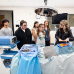 “A female healthcare professional is explaining a surgical training mannequin to a group of five students. The mannequin is covered in a gray sheet, revealing a simulated human torso with an orange representing an organ. Surgical instruments and equipment, including a ventilator, are visible on a nearby table. The setting appears to be a medical training room.”