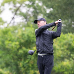 Man golfing outside wearing all black with trees in the background. It is raining.