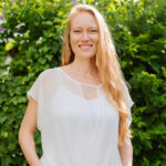 Female with long blonde hair stands outside in front of bushes. She is wearing a white top and pink pants