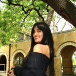 Female student with long dark hair wears an off the shoulder black top. She stands sideways and smiles at the camera. She is in front of a yellow building and trees
