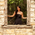 Female stands on other side of open windown with bricks around. She is wearing a black strapless dress with long dark hair