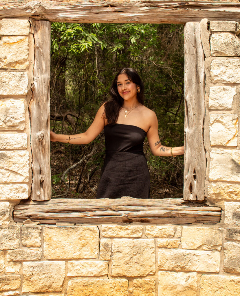 Female stands on other side of open windown with bricks around. She is wearing a black strapless dress with long dark hair 