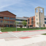 A modern educational building with a combination of brick and stone architecture. The structure features large windows and a prominent clock tower. In the foreground, there is a paved area with a stop sign and landscaped greenery, including small trees and grass. The sky is clear with a few clouds.