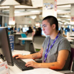 Student in grey shirt with purple lanyard works on computer. She is in a big computer lab with others working in the background