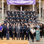 A large group photo of a diverse assembly of law enforcement officers and staff, standing on a staircase in a well-lit building. The officers are dressed in dark uniforms, while the staff members are in formal attire. In the foreground, a few individuals are standing in business suits, including two men in ties and a woman in a green dress. A banner above them reads "ACC is for Everyone." The setting appears to be a formal event or gathering.