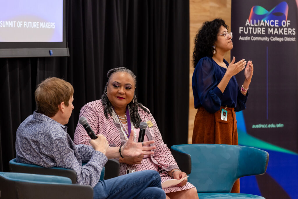 “Two women participate in a discussion at a summit. A Black woman with curly hair and a patterned shirt speaks into a microphone, gesturing with her hands. To her right, a young Black woman with curly hair wears a blue blouse and speaks with her hands raised. A third person, a white man, sits to the left, holding a microphone and looking at the speakers.”