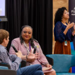 “Two women participate in a discussion at a summit. A Black woman with curly hair and a patterned shirt speaks into a microphone, gesturing with her hands. To her right, a young Black woman with curly hair wears a blue blouse and speaks with her hands raised. A third person, a white man, sits to the left, holding a microphone and looking at the speakers.”