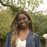 Student stands and smiles at the camera wearing graduation gown that is open with white top underneath. She is standing outside with trees in the background.