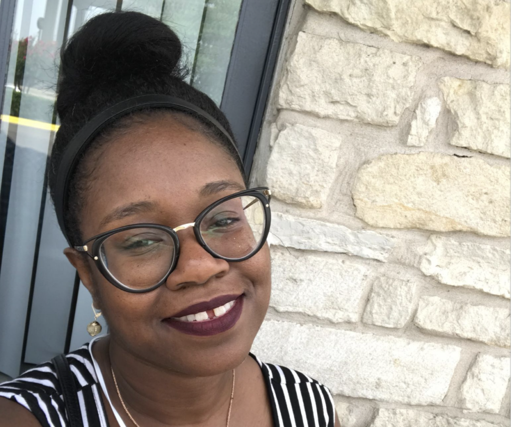 Black female smiles at the camera with her hair in a bun. She is wearing glasses and a black and white strap shirt. She is in front of a window and field stone wall