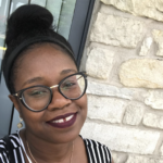 Black female smiles at the camera with her hair in a bun. She is wearing glasses and a black and white strap shirt. She is in front of a window and field stone wall