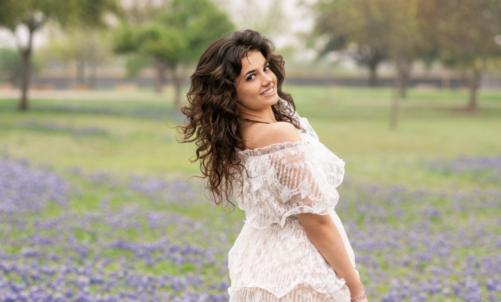 Girl with long brown hair and in white dress stands in field of wildflowers. She has her back to the camera and face turned to look with a smile