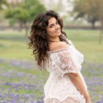 Girl with long brown hair and in white dress stands in field of wildflowers. She has her back to the camera and face turned to look with a smile