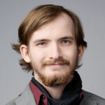 headshot of man with light brown hair. He is wearing a red collar shirt under a grey blazar. He has a beard