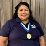 Female smiles at the camera wearing a gold medal. She is wearing a navy blue polo shirt in front of a wood paneled background