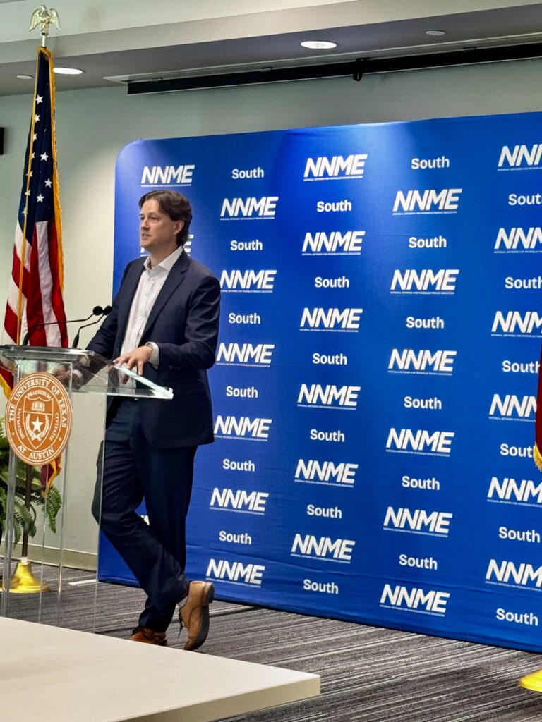 An image shows a medium shot of a man speaking at a clear podium with a seal from The University of Texas at Austin on the left. The man is wearing a dark blue suit, a white button-down shirt, and brown dress shoes. He is standing with his left leg crossed over his right leg, looking towards the left side of the frame. Behind him is a large blue banner with rows of text that read "NNME South." An American flag is on a flagpole to his left.