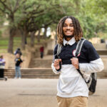 A young Black man with dreadlocks smiles at the camera while standing on a stone staircase on the Texas State Campus. He is wearing a black and white athletic shirt with a white collar, khaki pants, and carrying a gray backpack. Several other people are visible in the background, and trees surround the scene.