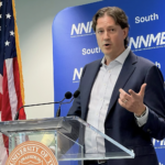 An image shows a medium shot of a man speaking at a clear podium with a seal from The University of Texas at Austin on the left. The man is wearing a dark blue suit, a white button-down shirt, and brown dress shoes. He is standing with his left leg crossed over his right leg, looking towards the left side of the frame. Behind him is a large blue banner with rows of text that read "NNME South." An American flag is on a flagpole to his left.