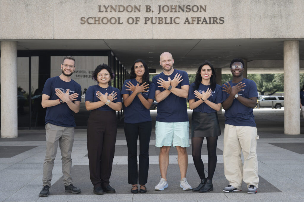 Six students stand outside the Lyndon B. Johnson School of Public Affairs building at the University of Texas, smiling with their arms crossed over their chests in a matching pose. They are wearing navy Texas LBJ School T-shirts and represent a diverse group. The building's name is prominently displayed above the entrance, and the group is centered in front of the school on a clear day.