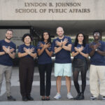 Six students stand outside the Lyndon B. Johnson School of Public Affairs building at the University of Texas, smiling with their arms crossed over their chests in a matching pose. They are wearing navy Texas LBJ School T-shirts and represent a diverse group. The building's name is prominently displayed above the entrance, and the group is centered in front of the school on a clear day.
