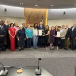 A group of approximately 18 people stands together in the Travis County Commissioners Court chamber for a recognition photo. Several people in the center hold framed proclamations while the group smiles toward the camera. The Travis County seal and U.S. and Texas flags are displayed behind them at the dais, with commissioners, community partners, and Austin Community College representatives gathered to celebrate the recognition.