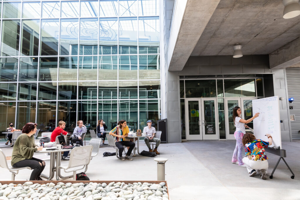 A group of students is engaged in a collaborative learning environment in an outdoor courtyard. Some students are seated at tables, working on laptops and discussing, while others are gathered around a whiteboard where one student is presenting. The setting features modern architecture with large glass windows and a concrete structure, creating a bright and open atmosphere. Pebbles are arranged in a decorative manner in the foreground.