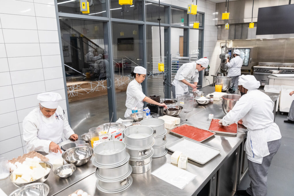Alt text: A modern kitchen with several culinary students in white chef uniforms and hats working at stainless steel tables. One student is mixing ingredients in a bowl, while another is pouring batter into cake pans. Various baking tools and ingredients, including bowls and measuring cups, are spread across the tables. Large windows provide natural light, and kitchen equipment is visible in the background.