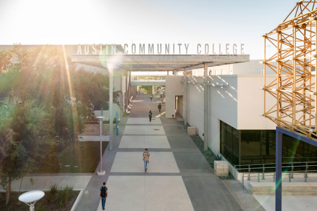 A sunny day at Austin Community College, featuring a wide pathway leading to the entrance of the campus. Students walk along the path, surrounded by modern architecture and greenery. A large sign reading "Austin Community College" is prominently displayed above the entrance, with a unique geometric structure visible on the right.