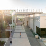 A sunny day at Austin Community College, featuring a wide pathway leading to the entrance of the campus. Students walk along the path, surrounded by modern architecture and greenery. A large sign reading "Austin Community College" is prominently displayed above the entrance, with a unique geometric structure visible on the right.