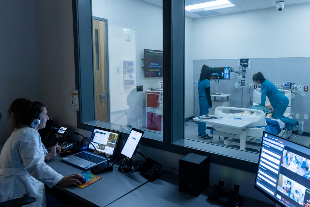 A medical simulation room is depicted, viewed through a large glass window. Inside, two healthcare professionals in scrubs are attending to a patient lying on a hospital bed. One is performing a procedure while the other assists. In the foreground, a person wearing a white lab coat and headphones is seated at a desk with multiple computer screens, monitoring the situation. The room is equipped with medical equipment and displays showing vital signs.