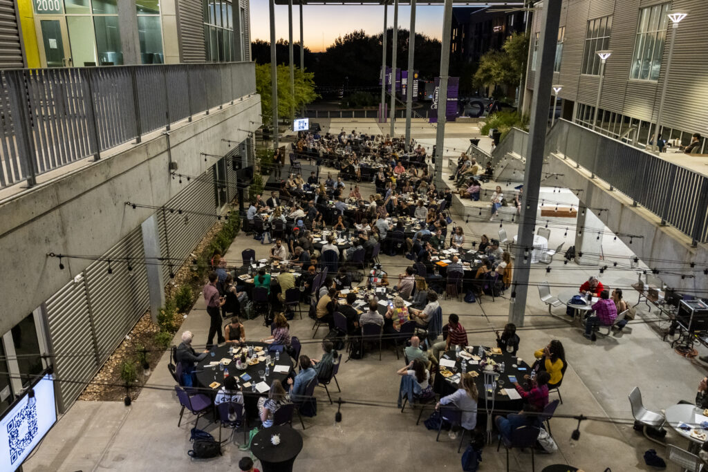 An aerial view of a large outdoor gathering in a modern courtyard. Numerous tables are filled with people engaged in conversation and dining. The setting features contemporary architecture with tall pillars and large windows. In the background, there are trees and a sunset sky, creating a warm atmosphere. Some people are using laptops, while others are enjoying food and drinks. The scene conveys a lively and social environment.