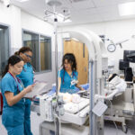 Three healthcare professionals in scrubs are gathered around a medical workstation in a hospital room. One nurse is holding a document and reviewing it, while the other two are focused on the equipment and a patient simulation model on a nearby bed. The room is well-lit and equipped with various medical devices, including monitors and IV stands.