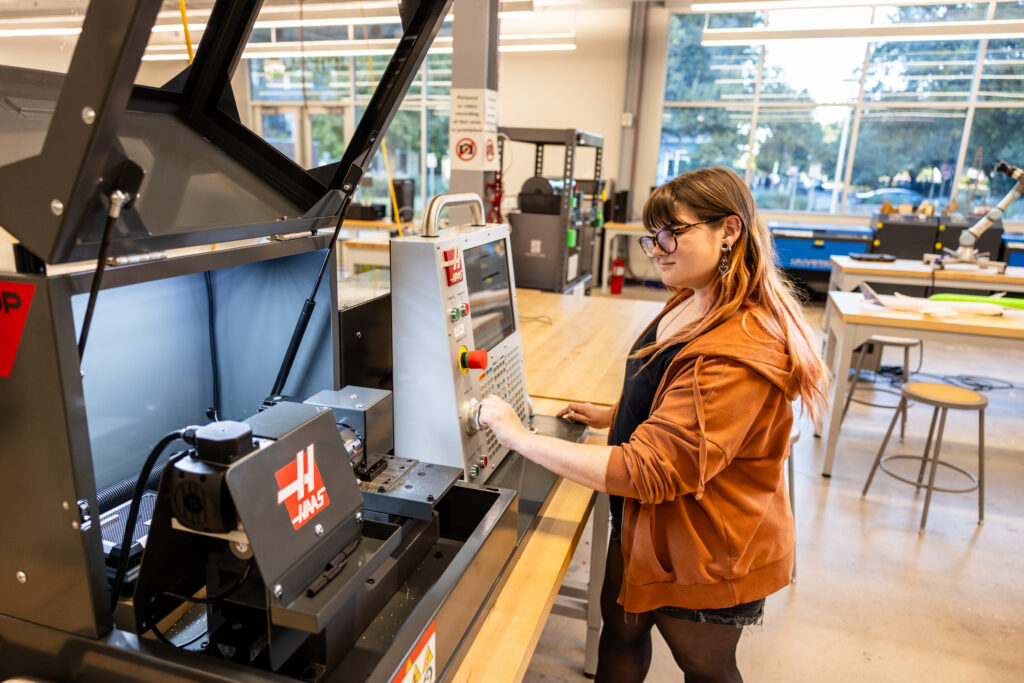 A young woman with glasses and long hair, wearing an orange hoodie, is operating a CNC machine in a workshop. She is focused on the control panel, which features various buttons and switches. The workshop is well-lit with large windows in the background, showcasing a bright and modern environment. Worktables and tools are visible in the space around her.
