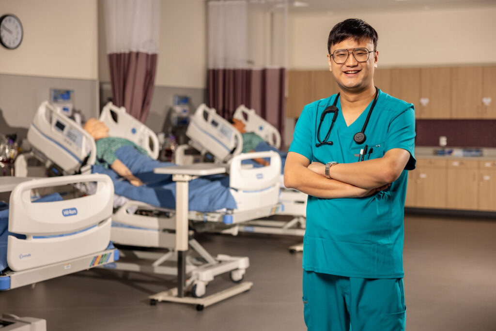 Here's an alt text description for the image: “A smiling Asian male healthcare professional wearing a teal scrubs uniform and stethoscope stands in a hospital room. He has glasses and his arms are crossed. In the background, there are two patient simulation mannequins lying on adjustable hospital beds, and medical equipment is visible.”