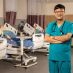 Here's an alt text description for the image: “A smiling Asian male healthcare professional wearing a teal scrubs uniform and stethoscope stands in a hospital room. He has glasses and his arms are crossed. In the background, there are two patient simulation mannequins lying on adjustable hospital beds, and medical equipment is visible.”