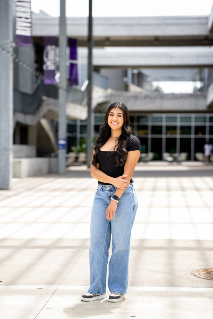 Here's an alt text description for the image: “A young woman with long, dark hair smiles directly at the camera while standing outdoors. She is wearing a black t-shirt and light-wash, wide-leg jeans. Her hands are crossed in front of her, and she is wearing a watch. The background is a modern, concrete plaza with a large building structure above, featuring purple signage.”
