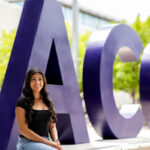 Here's an alt text description for the image: “A young woman with long, dark hair is seated outdoors in front of large, purple “ACC” letters. She is wearing a black top and jeans and is smiling at the camera. The background includes a bright, sunny sky and lush green trees.”