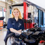 Here's an alt text description for the image: “A young woman wearing safety glasses and black work gloves stands beside an open engine bay of a vehicle in an automotive technology workshop. She is wearing a dark blue polo shirt with the logo “North Broward Tech.” The engine is a black inline-four engine with various components visible. The workshop is brightly lit and features red and blue overhead lighting, along with automotive tools and equipment in the background.”