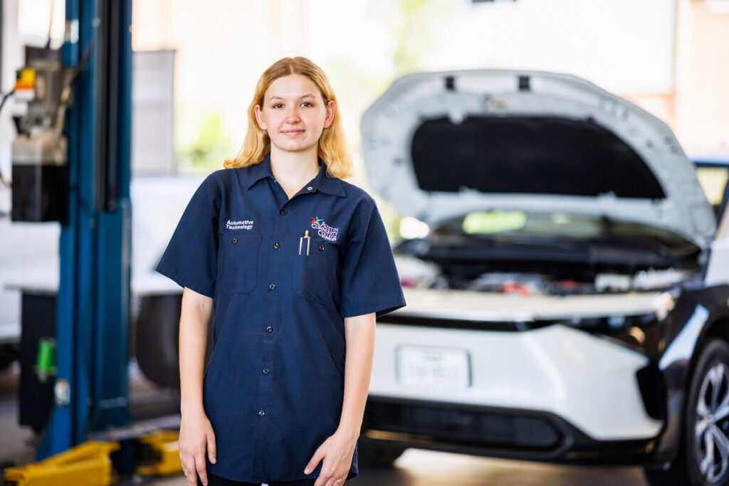 Here's an alt text description for the image: “A young woman wearing a dark blue mechanic’s shirt with a logo stands in front of an open hood of a white Honda Civic. A lift is visible in the background.”