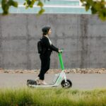 woman rides Lime scooter with white base and green handle. She is on a sidewalk in front of a grey wall