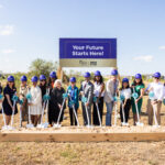 A group of diverse individuals stands in front of a large sign that reads "Your Future Starts Here!" They are gathered around a wooden platform filled with sand, each holding a shovel. Everyone is wearing blue hard hats and teal ribbons, symbolizing a groundbreaking ceremony. The background features a clear blue sky and some greenery.
