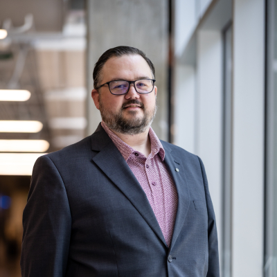 A man stands confidently near a large window in a modern office space. He has a beard and wears glasses, dressed in a dark blazer over a light-colored shirt. His hands are casually tucked into his pockets, and he is smiling slightly. The background features a blurred office environment with blue chairs and soft lighting.