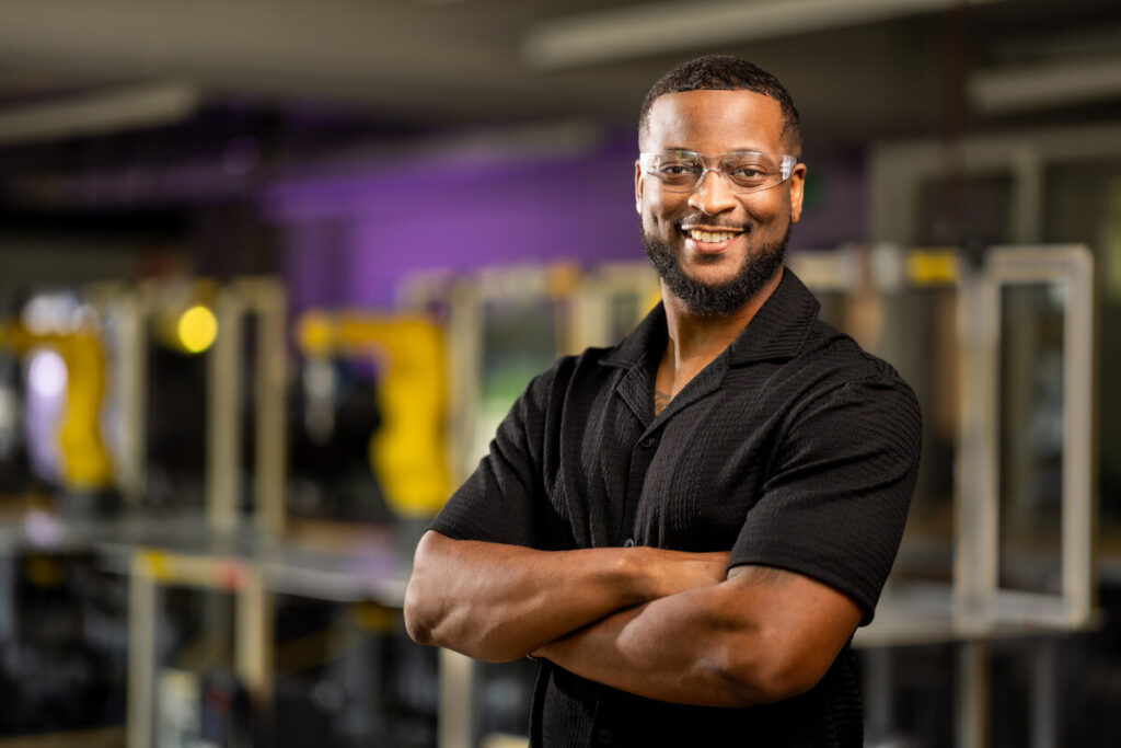 Here's an alt text description for the image: “A smiling Black man wearing glasses and a black button-down shirt stands with his arms crossed. He is in a workspace with several metal frames visible in the background, painted in shades of yellow and purple.”