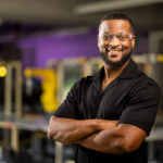 Here's an alt text description for the image: “A smiling Black man wearing glasses and a black button-down shirt stands with his arms crossed. He is in a workspace with several metal frames visible in the background, painted in shades of yellow and purple.”