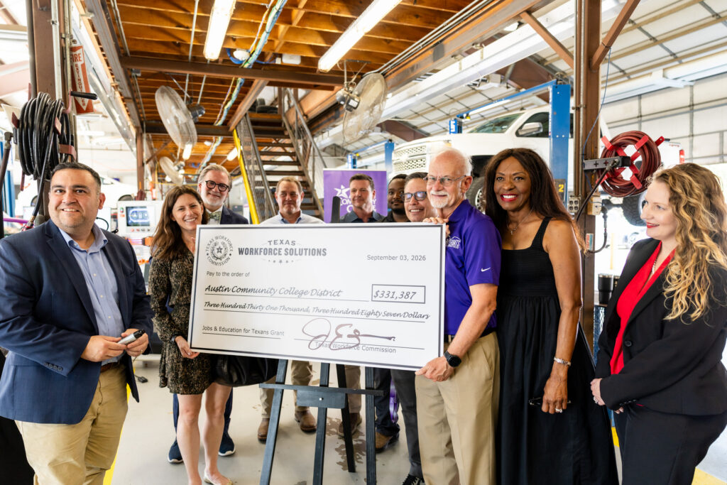 A group of people stands in a workshop setting, holding a large novelty check made out to the Austin Community College District for $331,887. The check is dated September 27, 2023, and is presented by Texas Workforce Solutions. The group includes men and women of diverse backgrounds, smiling and posing together. The workshop features various tools and equipment in the background, indicating a focus on vocational training or education.