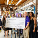 A group of people stands in a workshop setting, holding a large novelty check made out to the Austin Community College District for $331,887. The check is dated September 27, 2023, and is presented by Texas Workforce Solutions. The group includes men and women of diverse backgrounds, smiling and posing together. The workshop features various tools and equipment in the background, indicating a focus on vocational training or education.