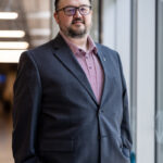 A man stands confidently near a large window in a modern office space. He has a beard and wears glasses, dressed in a dark blazer over a light-colored shirt. His hands are casually tucked into his pockets, and he is smiling slightly. The background features a blurred office environment with blue chairs and soft lighting.